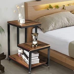 Mesa de noche de dos niveles con estructura metálica negra y superficies de madera oscura. Incluye un estante inferior para libros, una parte superior decorada con plantas y elementos decorativos.