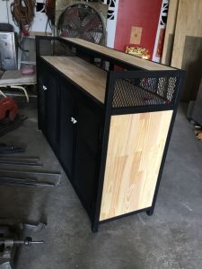 Mueble de almacenamiento rectangular con estructura de metal negro y paneles de madera clara. Tiene puertas de malla metálica y superficie lisa, ideal para espacios industriales o rústicos.