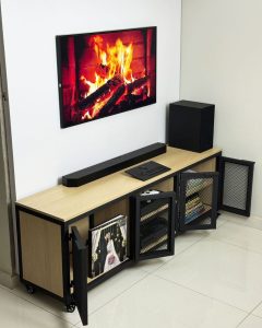 Mueble de TV moderno con superficie de madera clara y puertas de malla metálica negra. Incluye varios compartimentos para almacenamiento y ruedas para fácil movilidad.