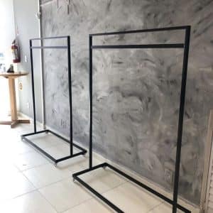 Dos percheros metálicos negros con estructura rectangular, sobre suelo de baldosas blancas y fondo de pared texturizada gris. Ideal para exhibición de ropa en tiendas o vestidores.