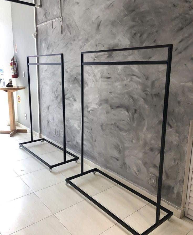 Dos percheros metálicos negros con estructura rectangular, sobre suelo de baldosas blancas y fondo de pared texturizada gris. Ideal para exhibición de ropa en tiendas o vestidores.