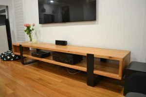 Mueble bajo de madera clara con diseño moderno, cuenta con estantes abiertos y patas de metal negro, ideal para colocar equipos electrónicos en una sala de estar.