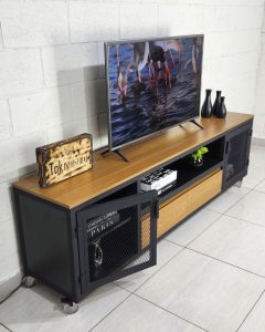 Mueble de televisión industrial con acabados de madera y negro, puertas de rejilla metálica y ruedas. Incluye estantes para almacenamiento. Diseño moderno y funcional.