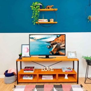 Mueble de TV de madera con estante superior donde se apoya un televisor y decoración, incluyendo cuadros y libros. Cuenta con tres nichos inferiores para almacenamiento.