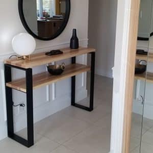 Mesa consola de madera clara con estructura metálica negra, diseñada para pasillos. Incluye dos estantes y está decorada con una lámpara y jarrones modernos.