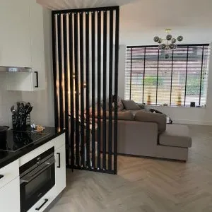 Panel divisor de madera negra con listones verticales, separando cocina y sala. Añade estilo moderno y funcionalidad a espacio abierto con decoración neutra.