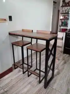 Mesa alta de estilo industrial con tablero de madera y estructura de metal negro, acompañada de dos taburetes a juego. Ideal para espacios pequeños o cocina moderna.