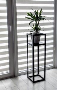 Soporte minimalista negro de metal alto con diseño cuadrado, ideal para exhibir plantas en interiores modernos, frente a persianas rayadas en un ambiente luminoso.