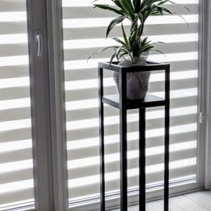 Soporte minimalista negro de metal alto con diseño cuadrado, ideal para exhibir plantas en interiores modernos, frente a persianas rayadas en un ambiente luminoso.