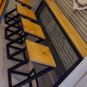 Cuatro taburetes de madera clara con estructura metálica negra y un diseño angular junto a una barra a juego en un entorno interior.