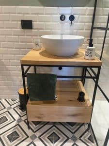 Mueble de baño minimalista con estructura metálica negra y encimera de madera clara. Incluye un lavabo de cerámica blanco y espacio de almacenamiento inferior con cajón y toalla verde.