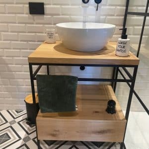Mueble de baño minimalista con estructura metálica negra y encimera de madera clara. Incluye un lavabo de cerámica blanco y espacio de almacenamiento inferior con cajón y toalla verde.