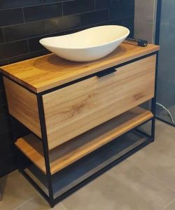 Mueble de baño moderno de madera con estructura metálica negra, lavabo blanco con diseño ovalado. Dispone de superficie amplia y repisa inferior para almacenamiento adicional.